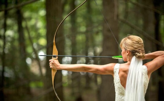 camp oneka weddings & gatherings bride in the woods aiming bow and arrow